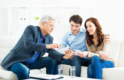 A man and a woman sitting on a couch, with the man's arm around the woman's shoulders. They are facing a 3rd white haired man who is showing them some paperwork