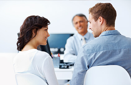 A man and a woman seated with their backs to the camera, with their head's turned to face each other, smiling. A man blurry in the background is seated behind a desk facing them