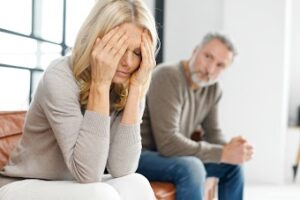 woman seated holding her head in her hands, with a man beside her looking at her concerned